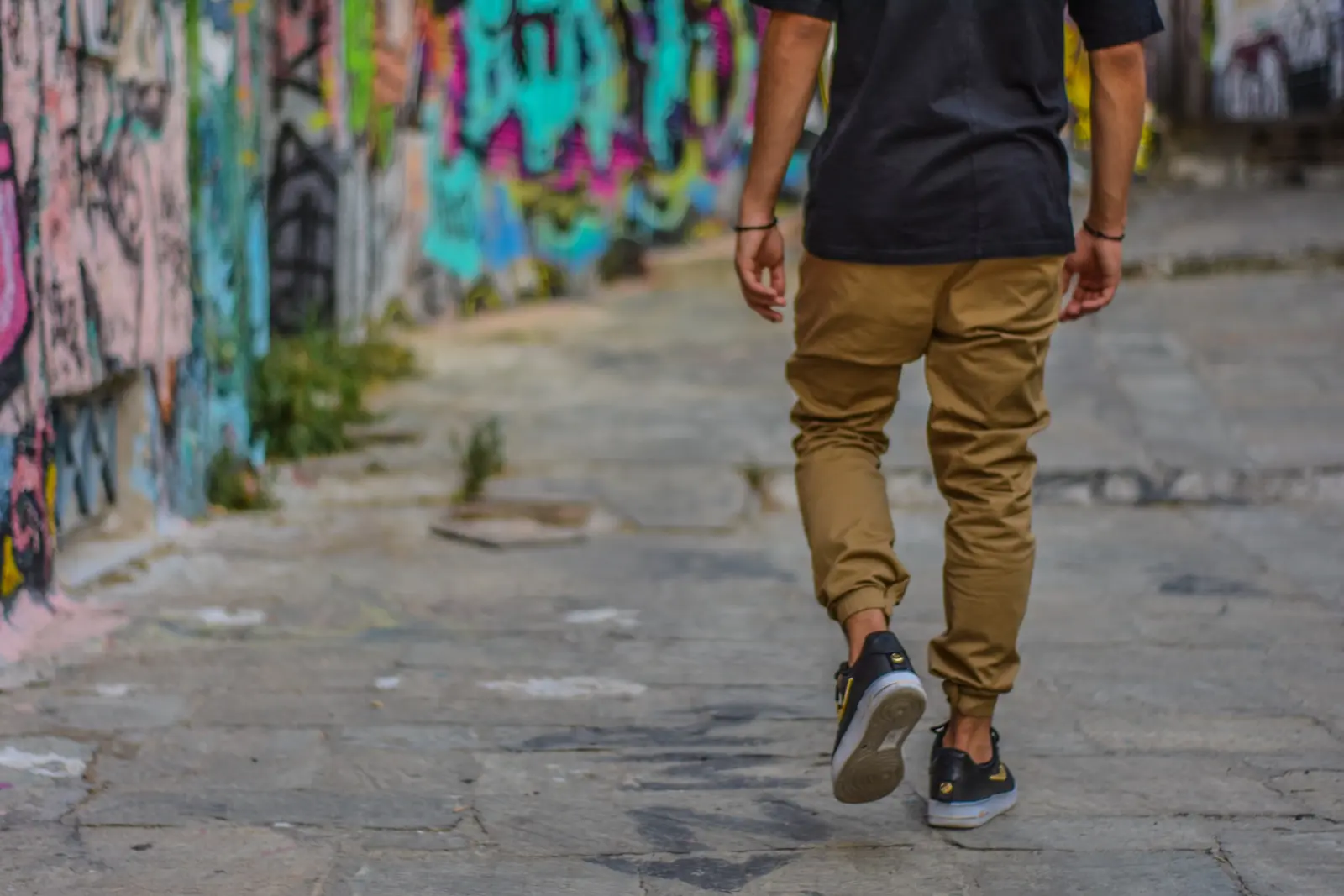 man wearing black shirt and brown men's pants walking beside wall with graffiti